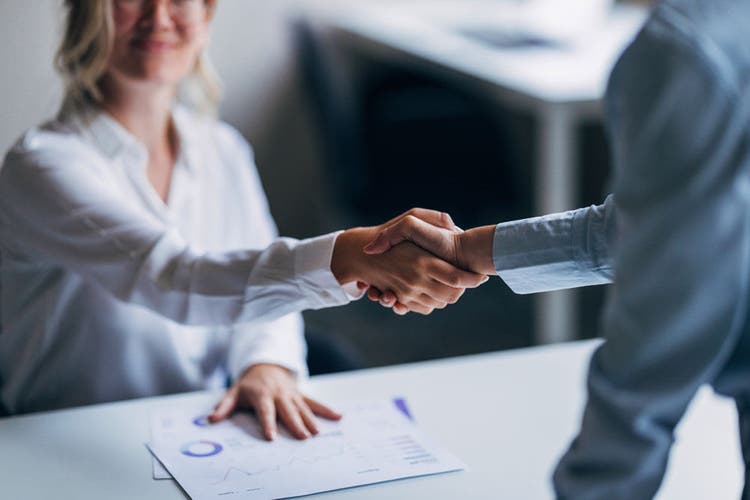 Two businesswomen confidently engage in a handshake at the office, symbolizing successful partnership and collaboration. Business documents are visible in the background.
