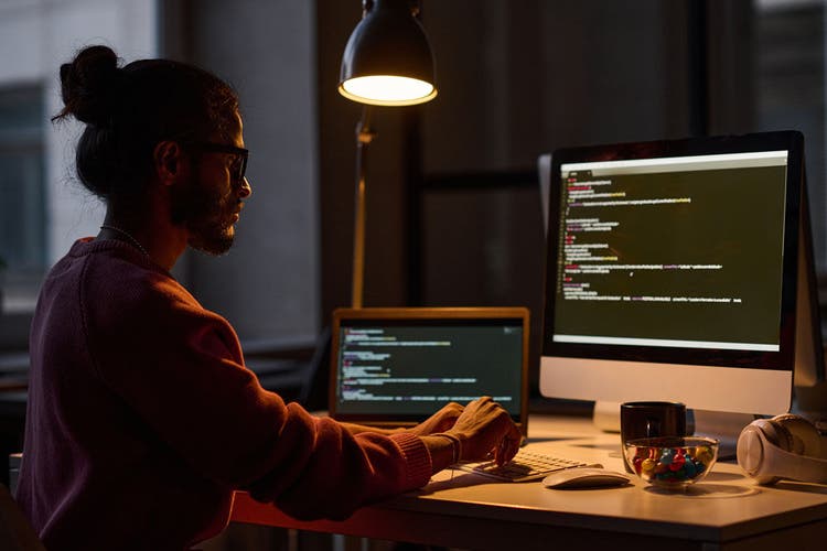 Young programmer sitting at his workplace and working on computer with codes, he working in office till late night