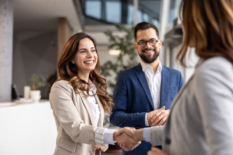 Two businesswomen and a businessman shaking hands in an office lobby, smiling and radiating happiness after successfully closing a lucrative deal, embodying teamwork and collaboration